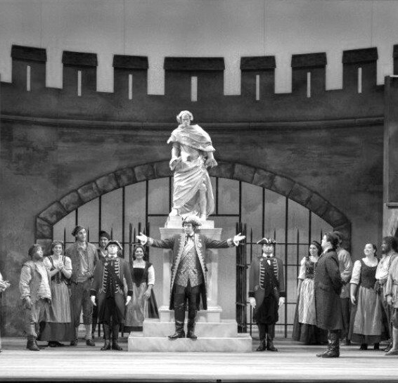 Stage scene with performers; statue on a pedestal, man gesturing; others in period costumes.