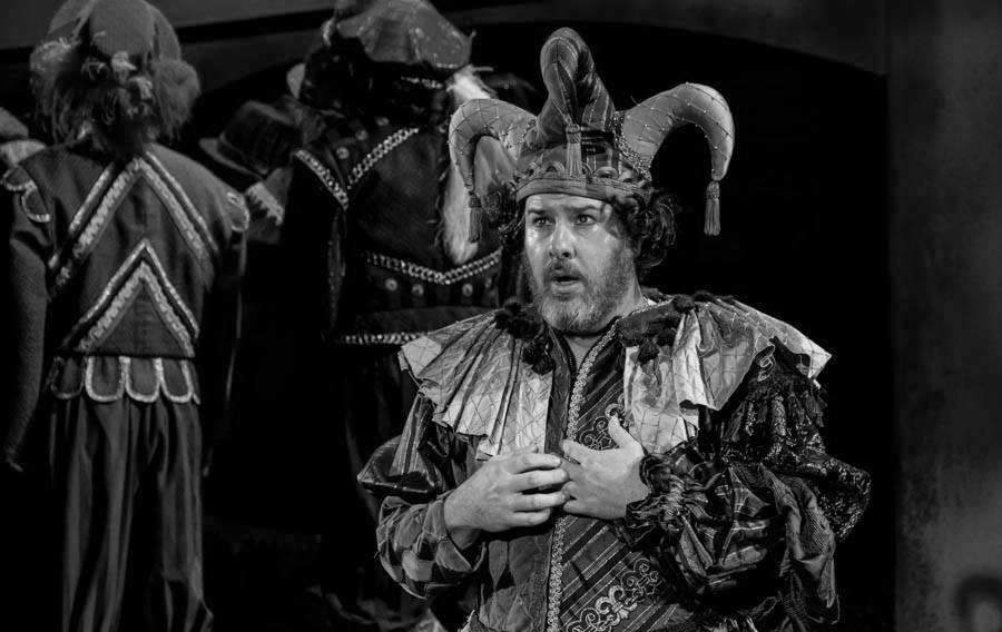 Man in jester costume on stage, hands on chest, other actors in background.