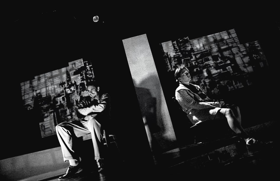 Two people seated in dark room, cityscapes projected behind them, a bubble floating above.