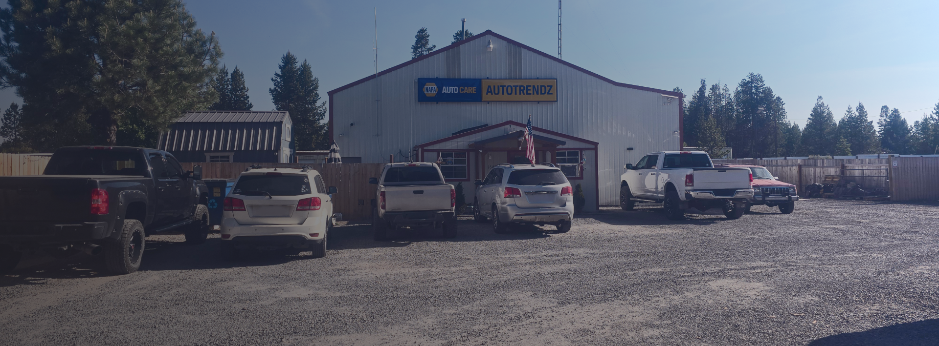 A white building with a blue sign, several parked cars, and an American flag on a gravel lot | Autotrendz