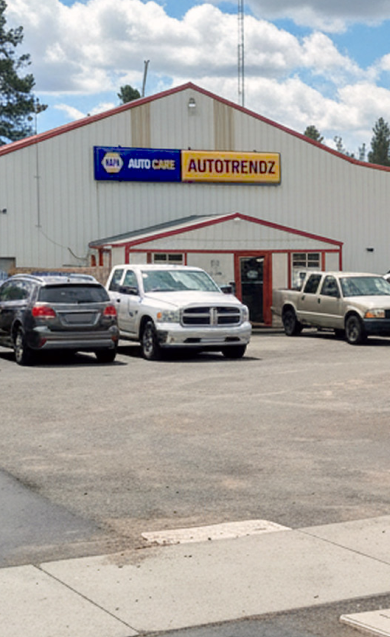 A white building with a blue sign, several parked cars, and an American flag on a gravel lot | Autotrendz