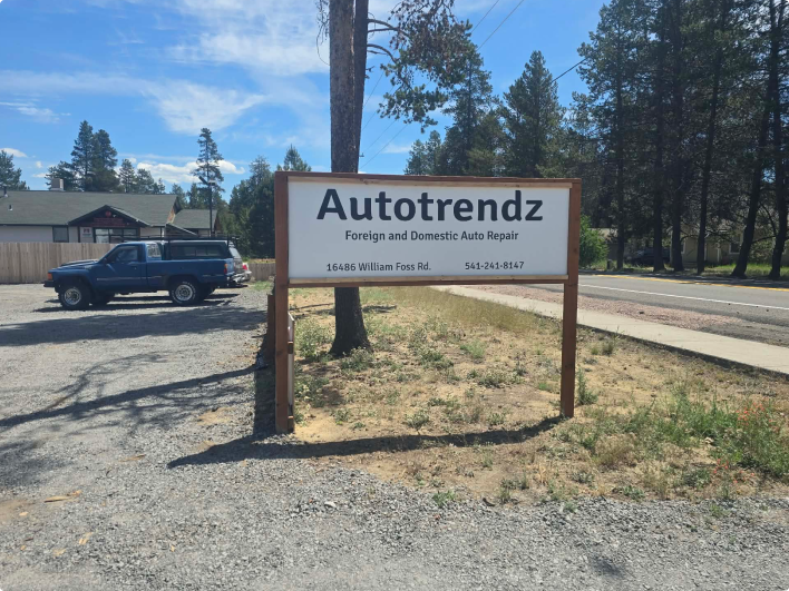 Autotrendz roadside sign beside a blue pickup, with trees and a road in the background | Autotrendz