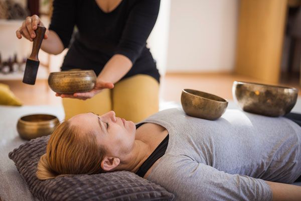 A woman is laying on a bed while a woman plays singing bowls on her back.