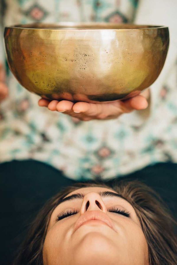 A woman is laying down with her eyes closed while a person holds a bowl over her head.