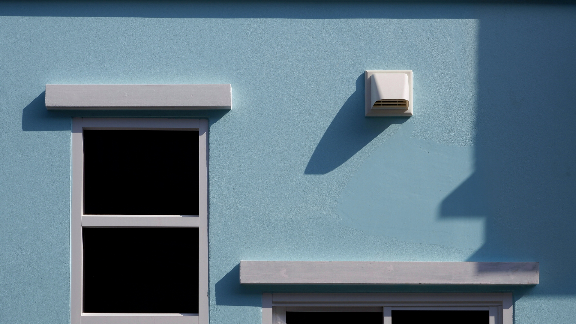 Blue stucco wall with white window trim and a ventilation hood casting a shadow.