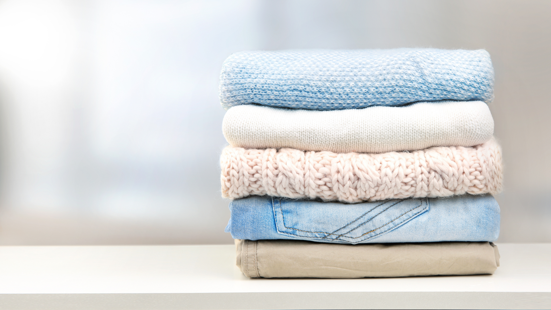 A stack of five folded garments in shades of blue, white, pink, and tan sitting on a white surface.