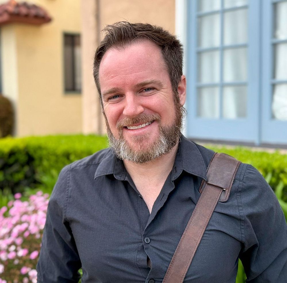 A man with a beard is sitting in a chair in front of a window.