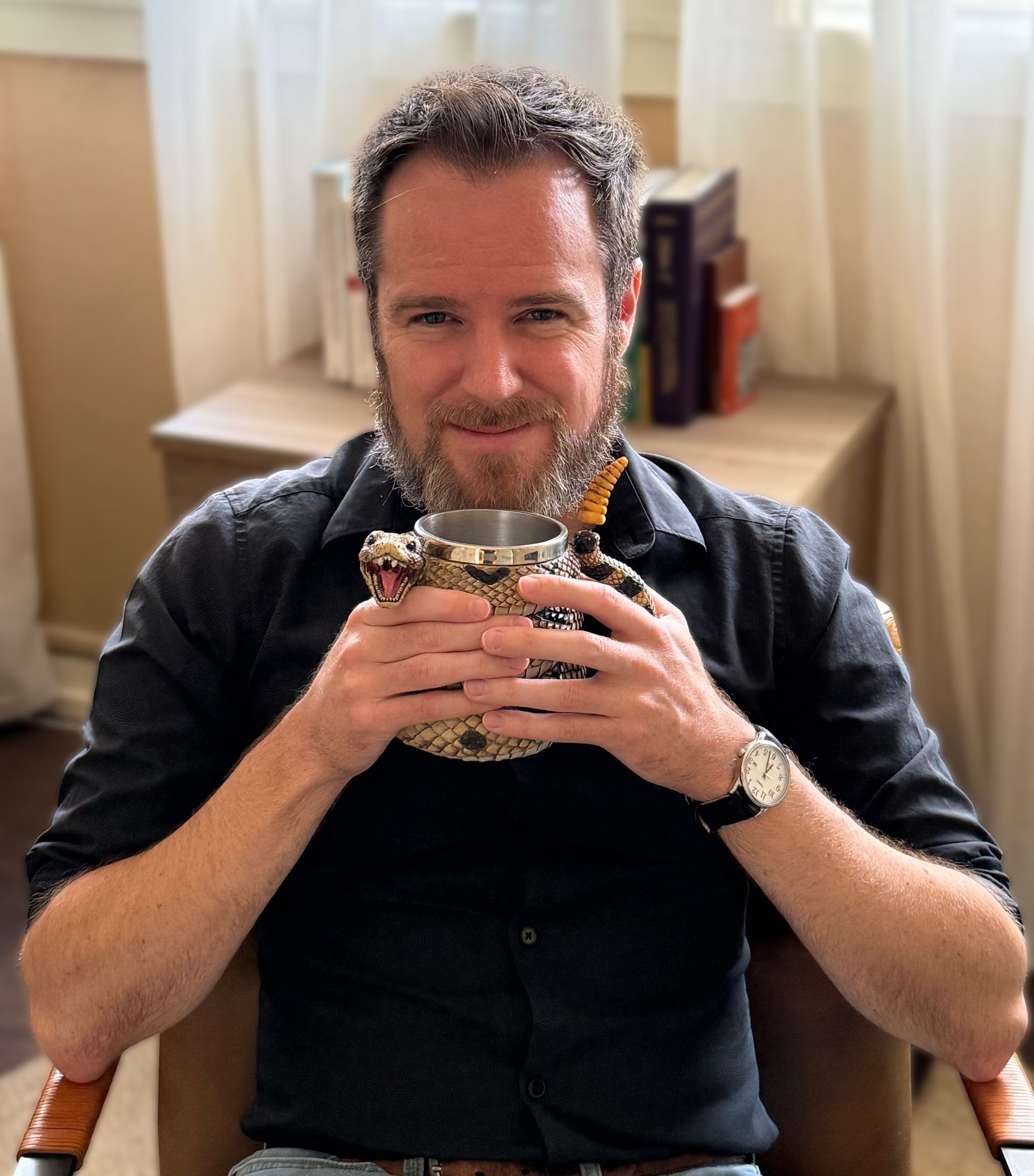 A man with a beard is sitting in a chair in front of a window.