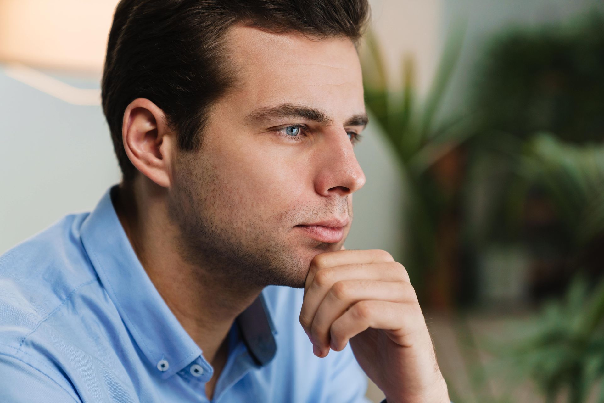A man in a blue shirt is sitting on a couch with his hand on his chin.