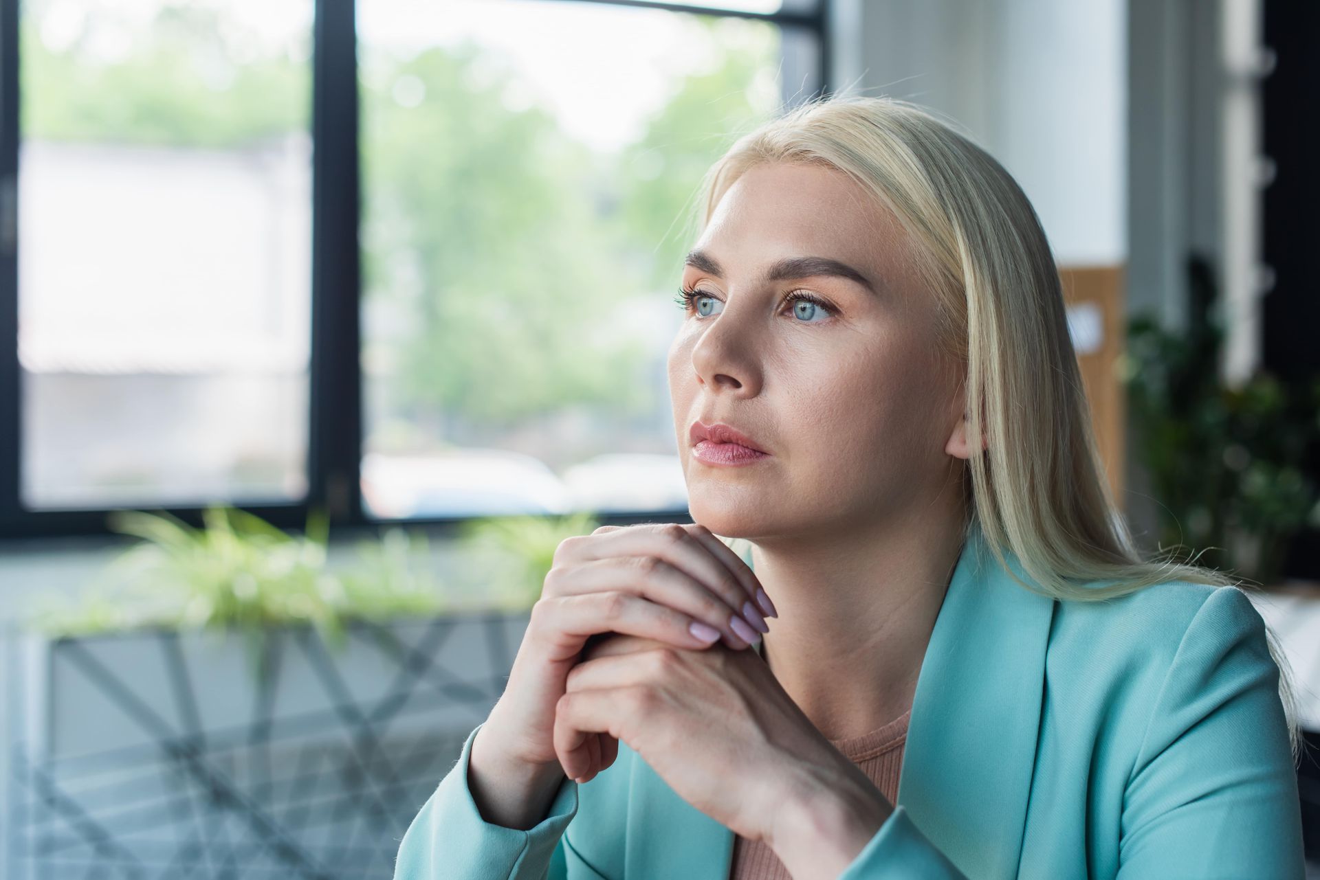 A woman is sitting at a table with her hands folded and looking out the window.