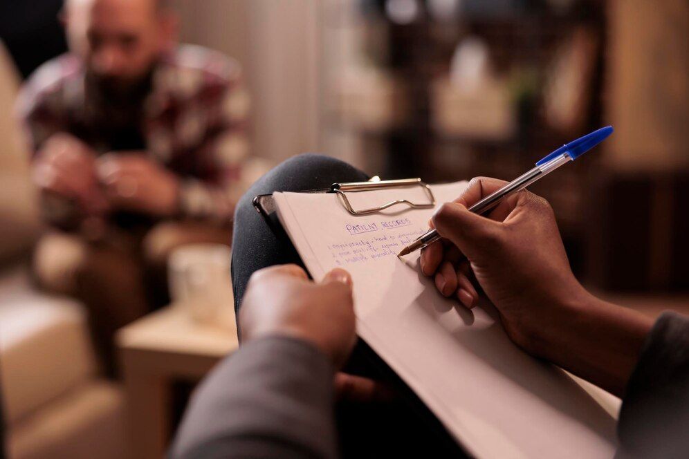 A person is writing on a clipboard while a man sits in the background.