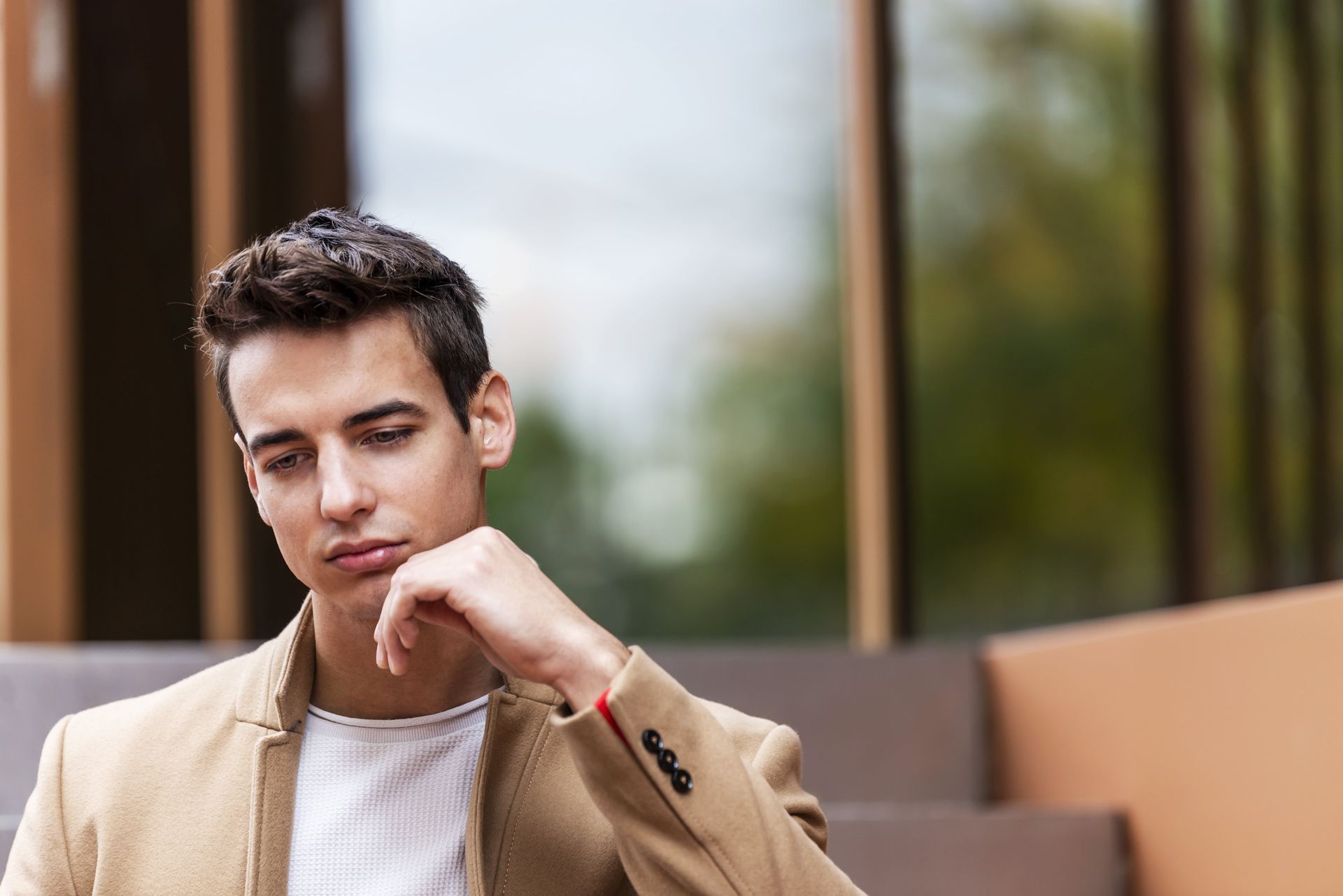 A young man in a tan jacket is sitting on a bench with his hand on his chin.