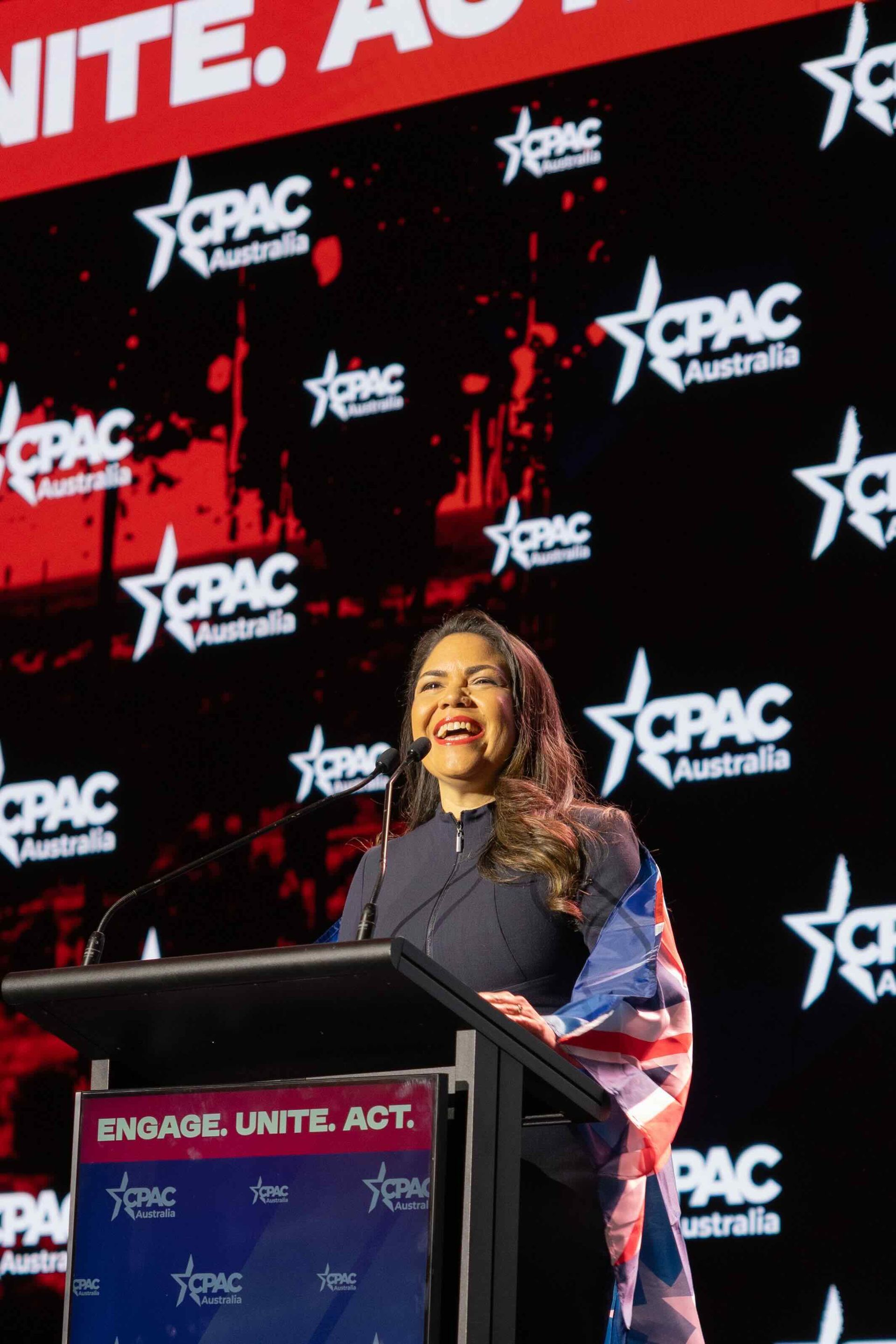 A woman at a podium with a flag draped on her shoulder, speaking at CPAC Australia.