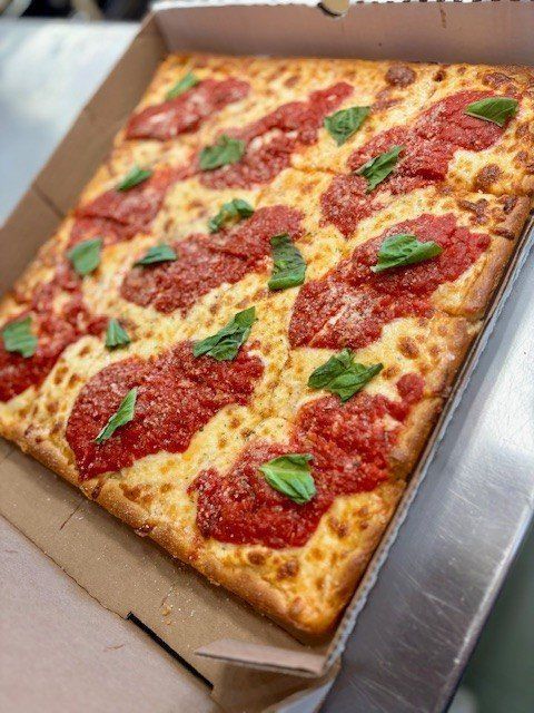 Square pizza with red sauce, cheese, and basil in a cardboard box.