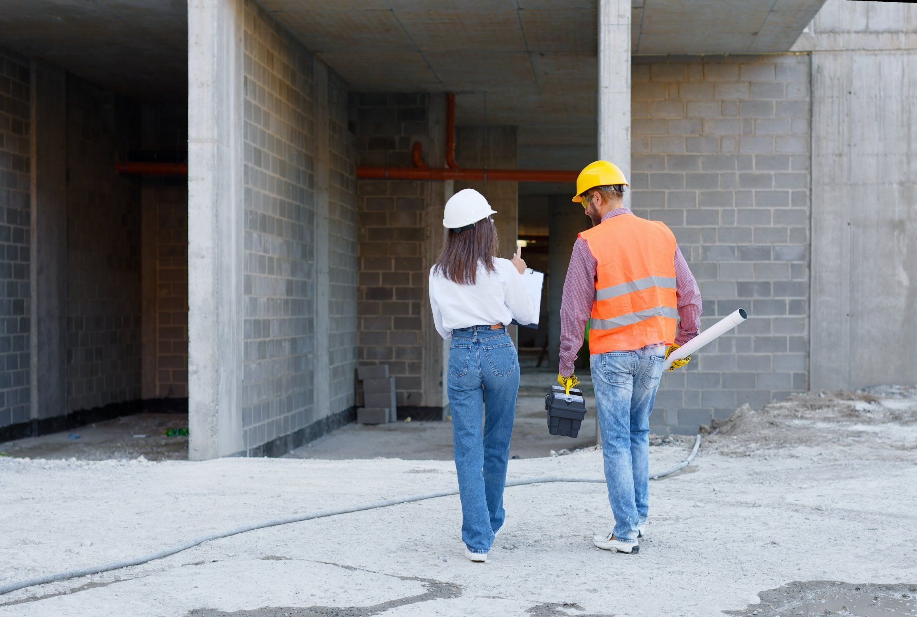 Two people, one in hard hat and vest, the other in white helmet, walking in a construction site, discussing plans.