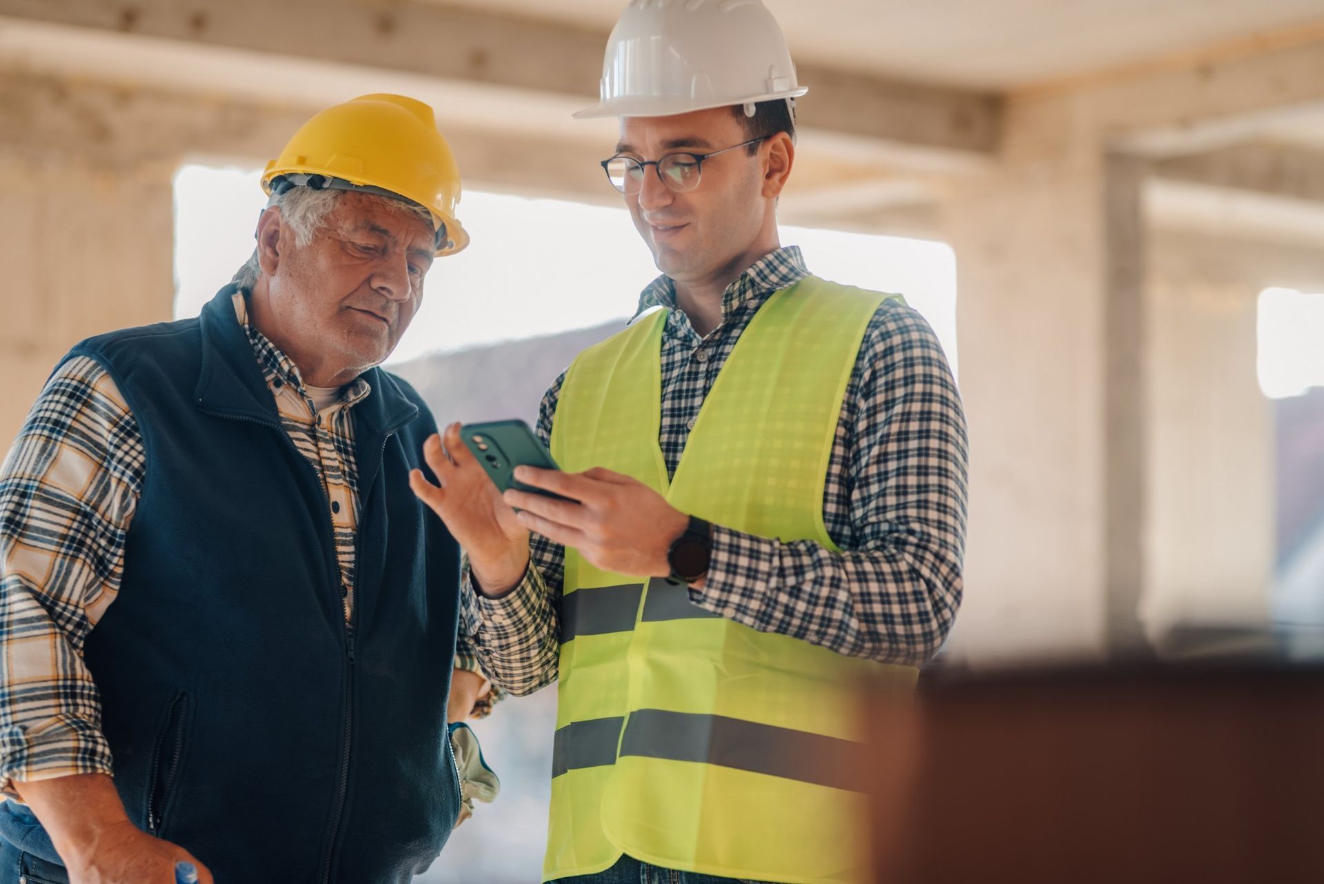 Two construction workers in hard hats looking at a phone on a construction site. One points.