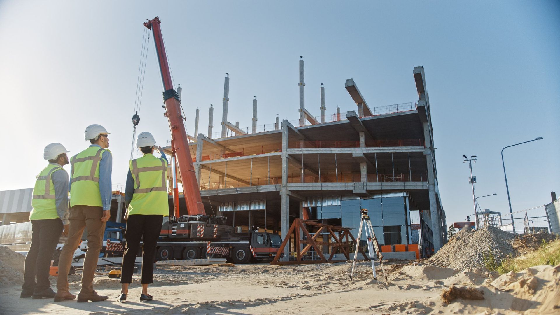 Construction workers in hard hats and vests observe a building under construction, a crane in the background.