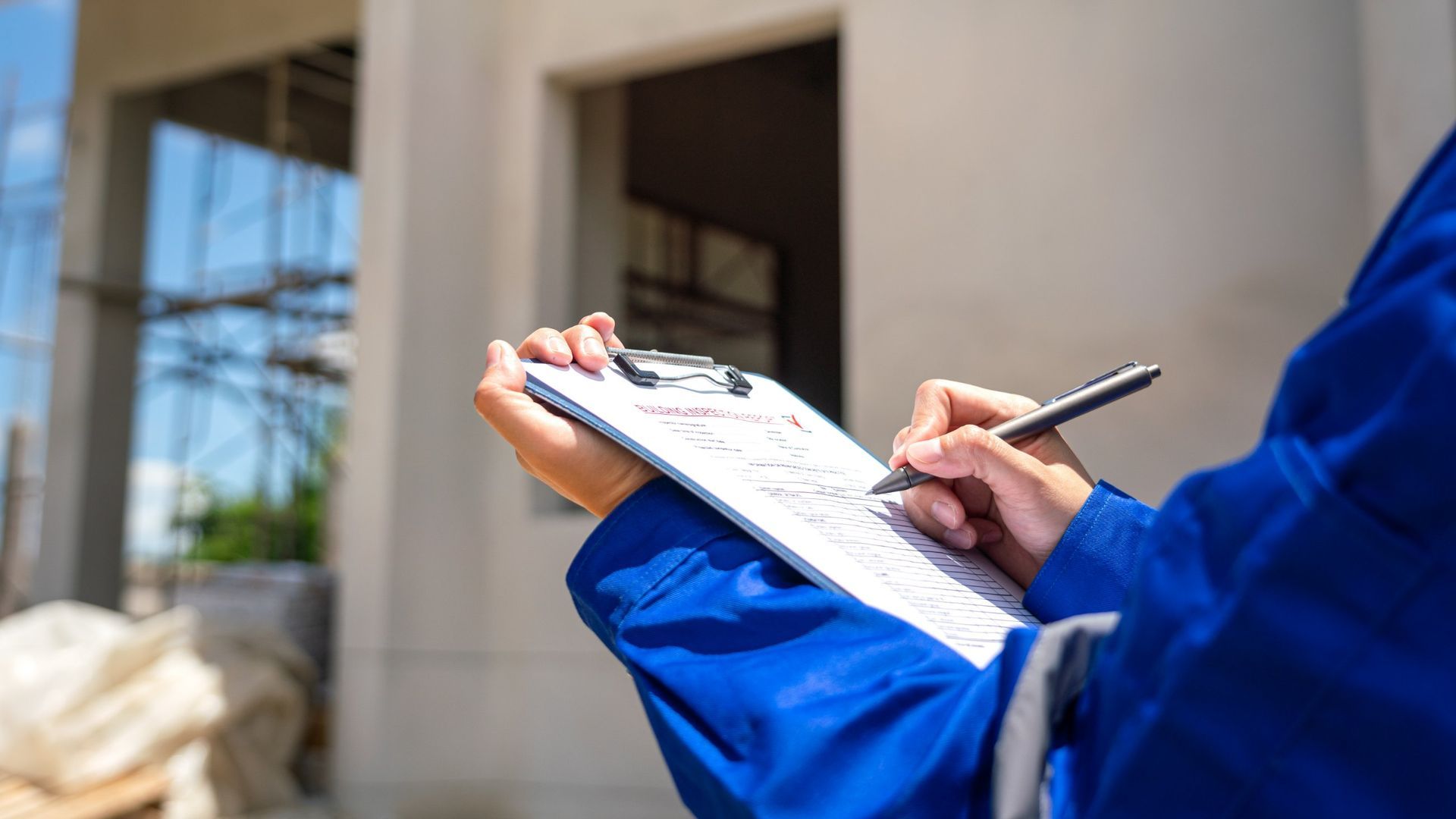 Person in blue jumpsuit taking notes on a clipboard at a construction site.