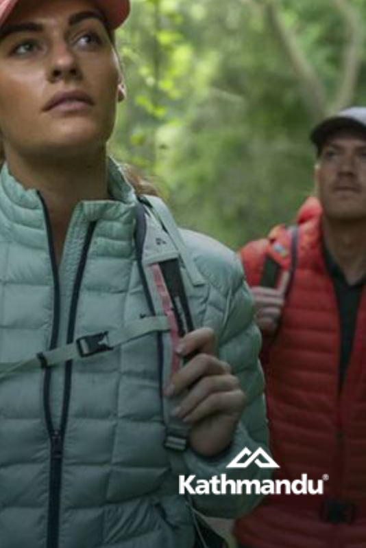 Woman wearing mint-green jacket and backpack looks upwards in a forest, Kathmandu logo at the bottom.