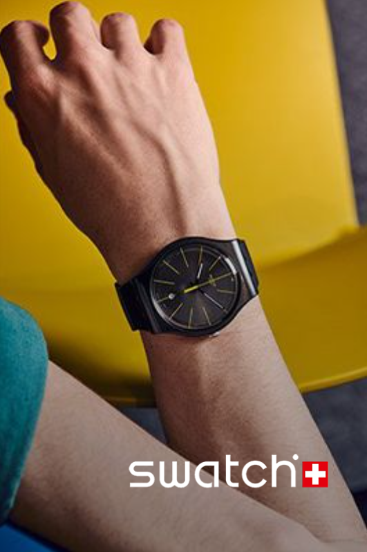 Black Swatch watch on wrist, yellow hands, against yellow chair.