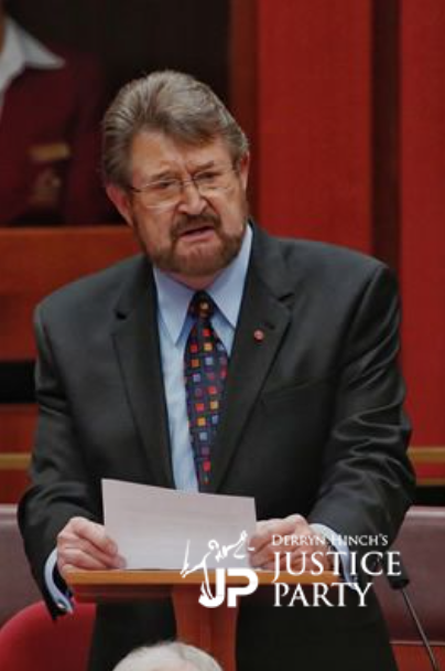 Man in suit speaking at a podium in a legislative chamber, Justice Party logo in foreground.