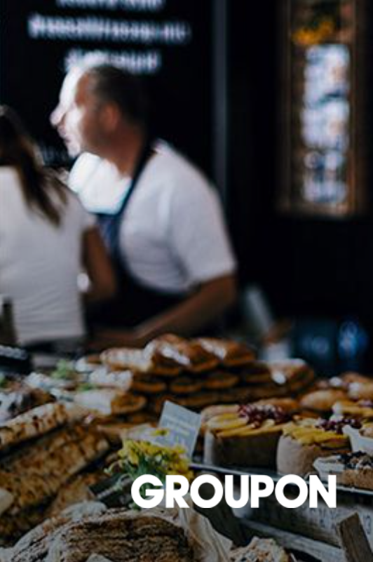 Groupon logo on a blurred background of baked goods and a person at a counter.