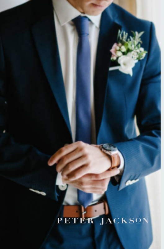 Man in navy blue suit, adjusting wristwatch. White shirt, blue tie, floral boutonniere, brown belt.
