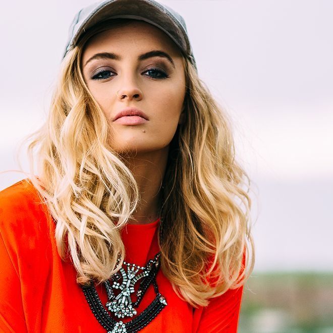 Woman with blonde wavy hair, wearing a baseball cap, orange shirt, and statement necklace; outdoors.