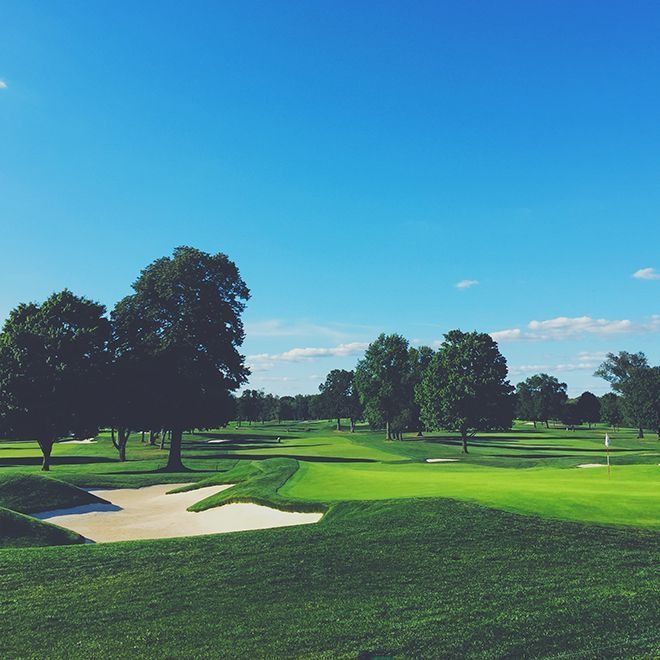 Green golf course with trees, sand trap, and blue sky.