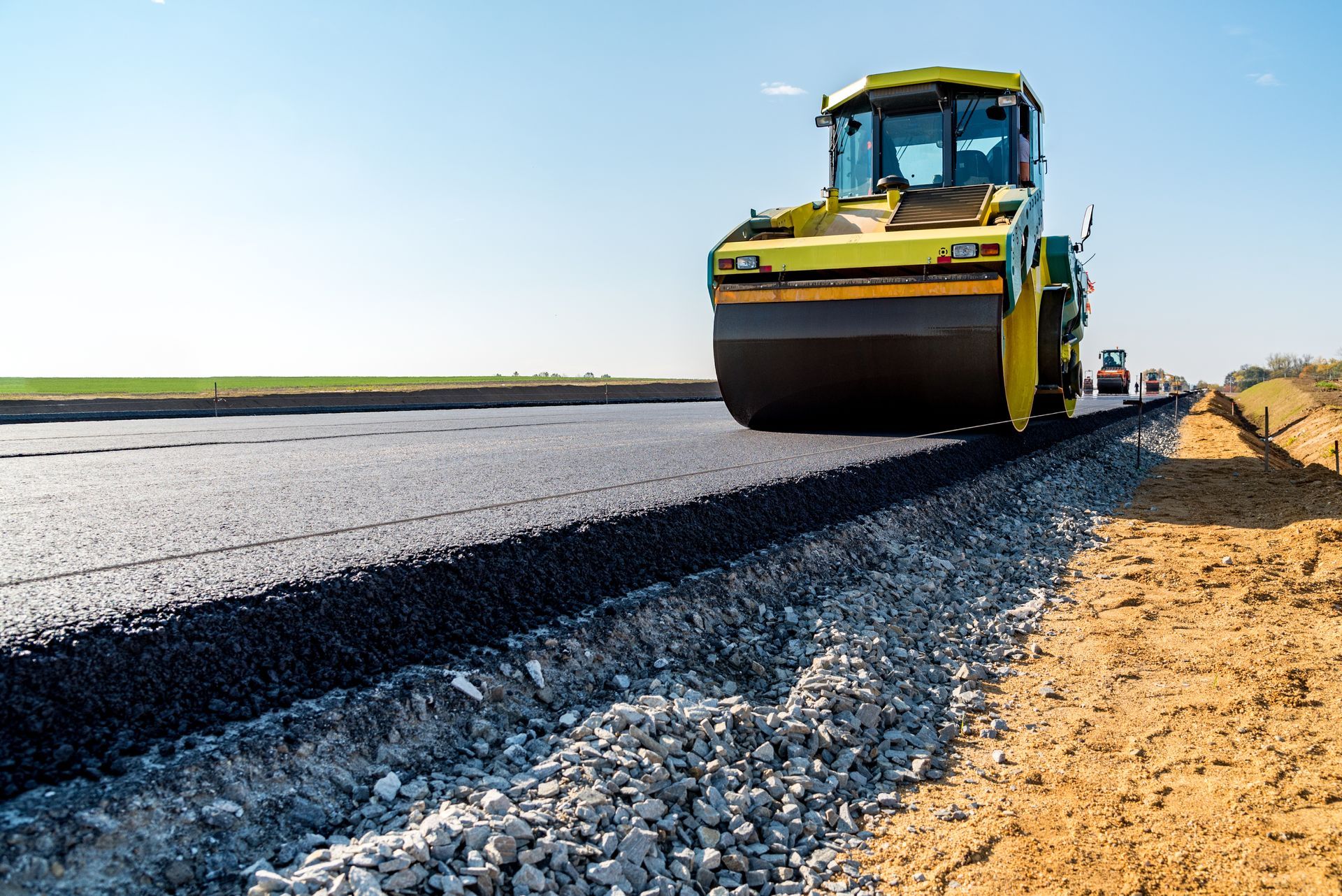 Road roller paving a new black asphalt road beside a sandy shoulder under a clear blue sky