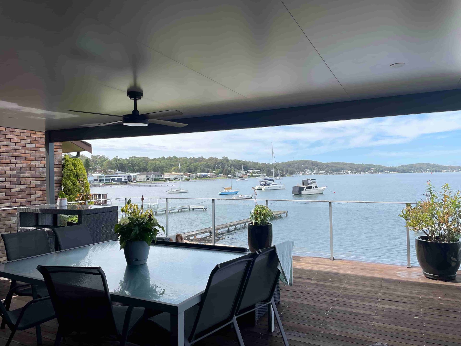 Outdoor deck overlooking water with boats. Table and chairs under a dark ceiling, with greenery.