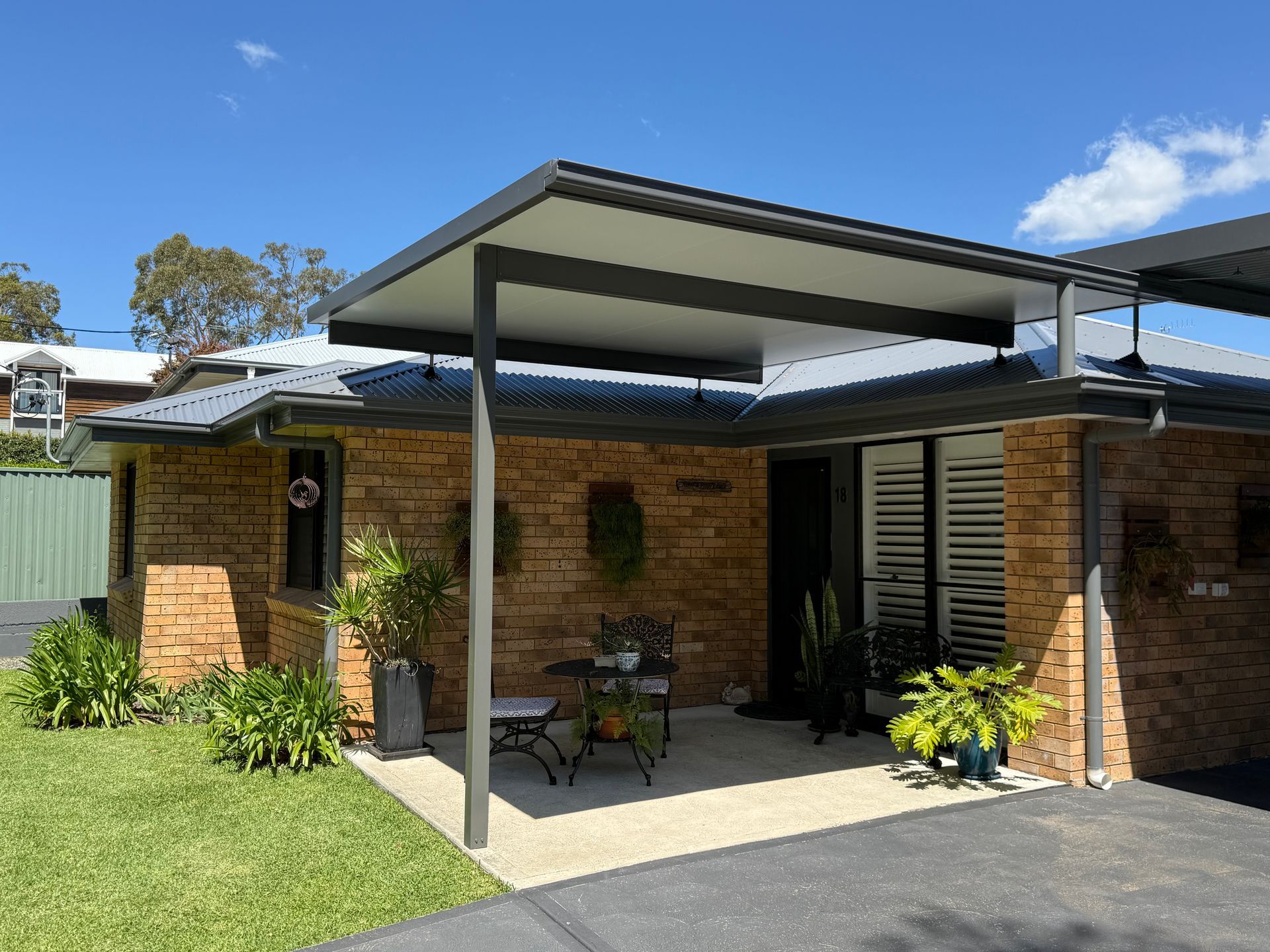 Brick house with covered patio, plants, and seating under a blue sky.