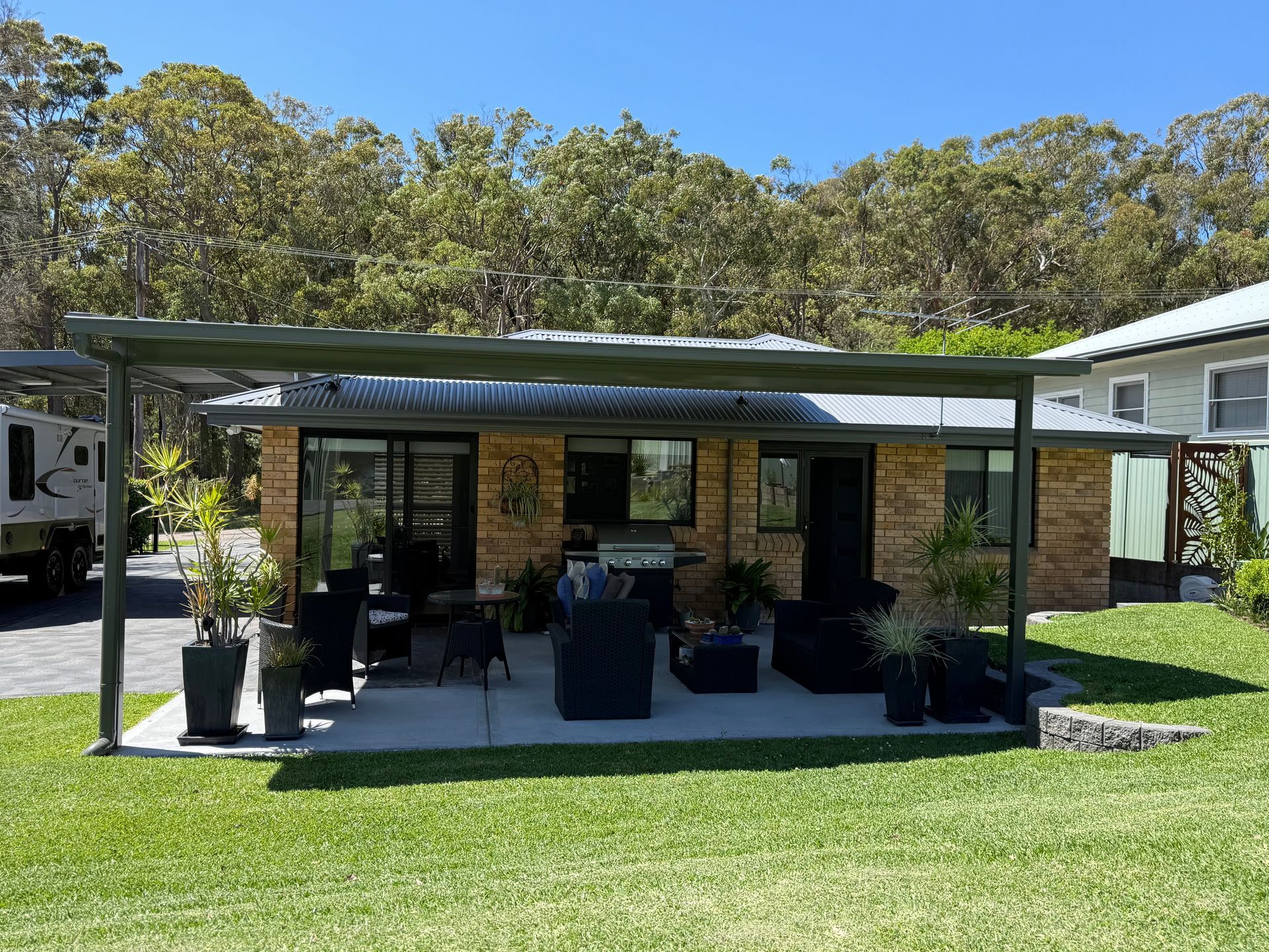 Brick house with patio and barbecue under a wooden pergola, set on a green lawn with trees in the background.