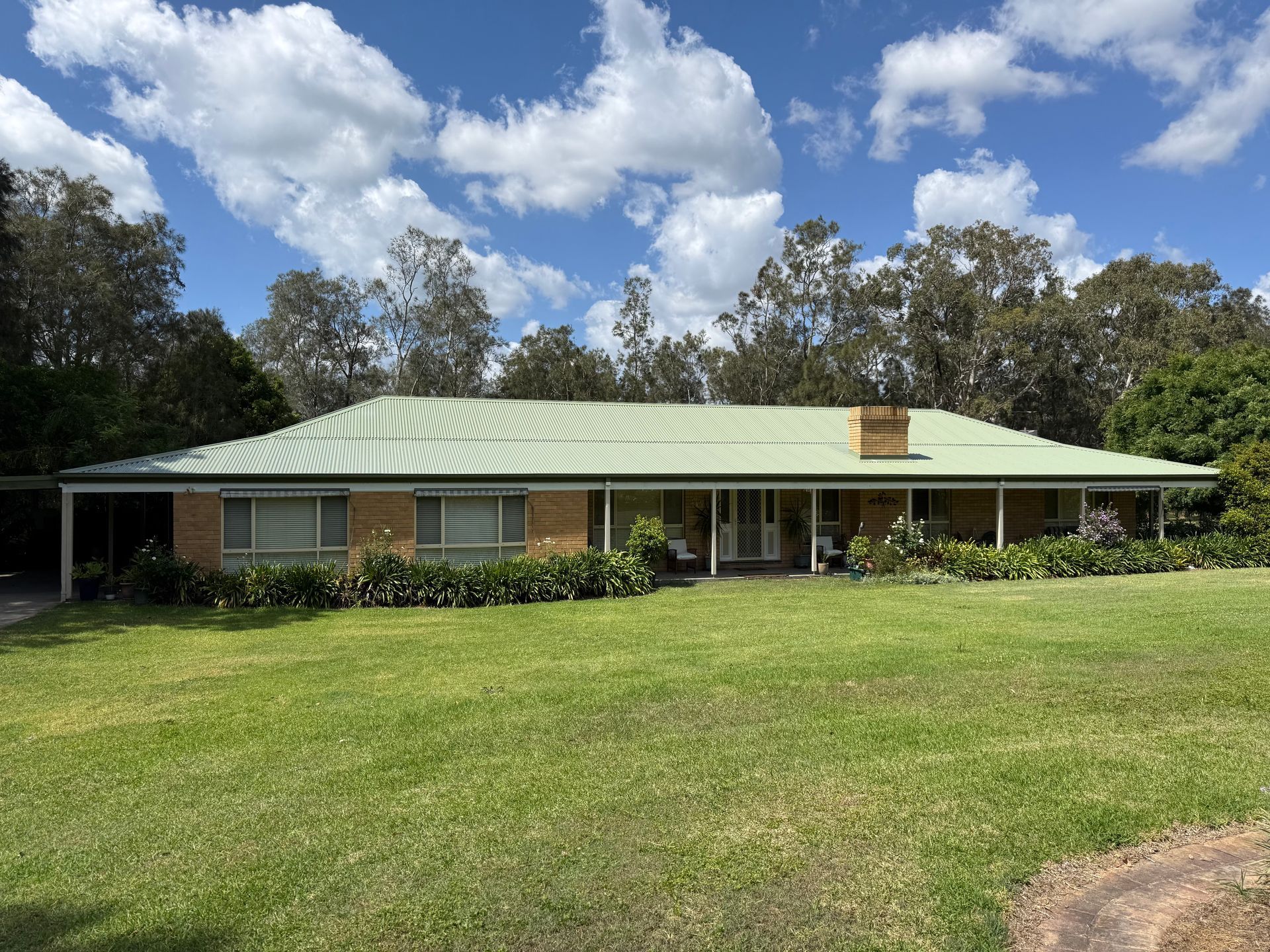 Single-story brick house with green roof, large lawn, and trees under a cloudy blue sky.
