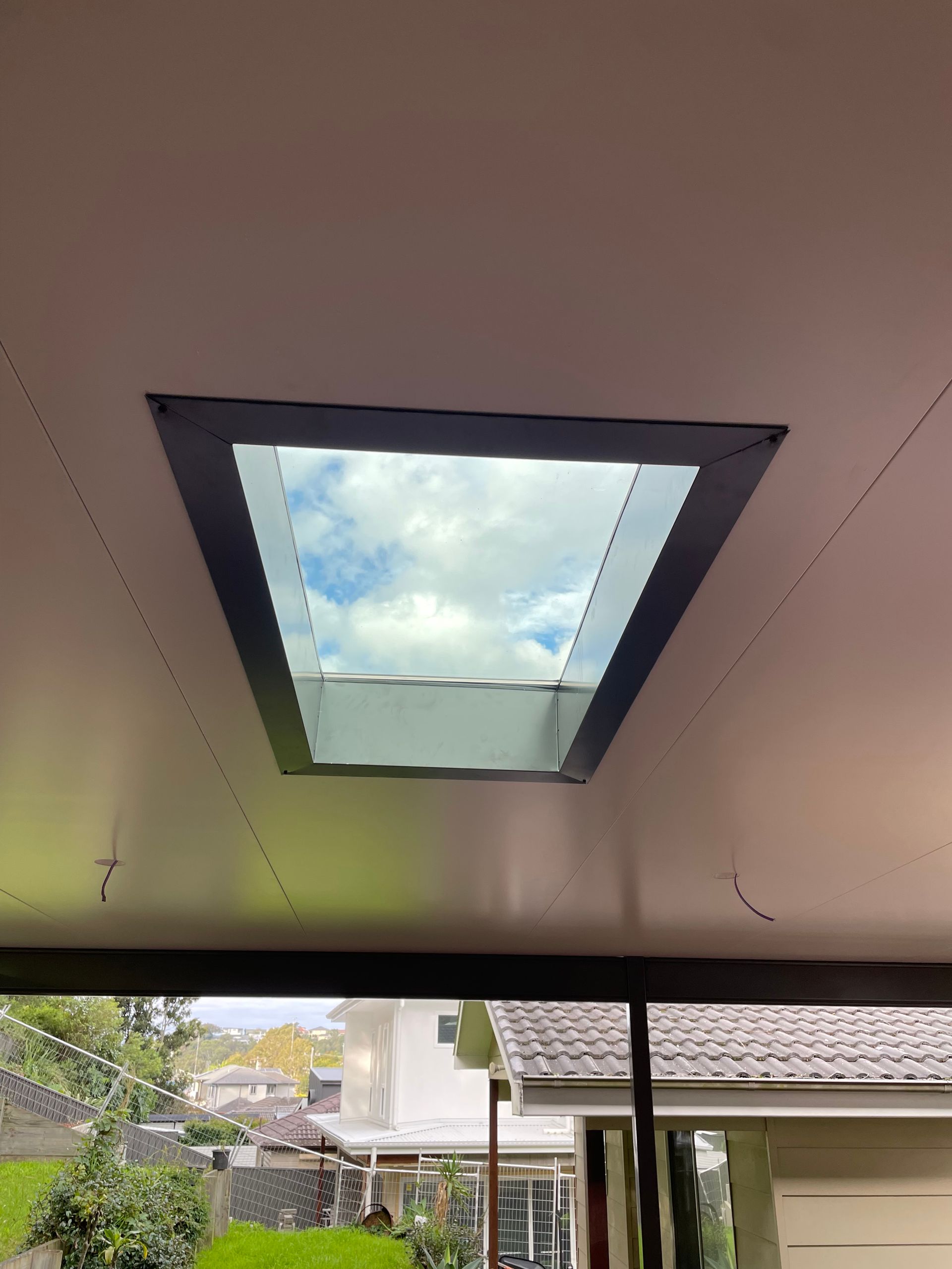 Skylight in a white ceiling, showing blue sky and clouds; exterior view of a backyard with greenery.