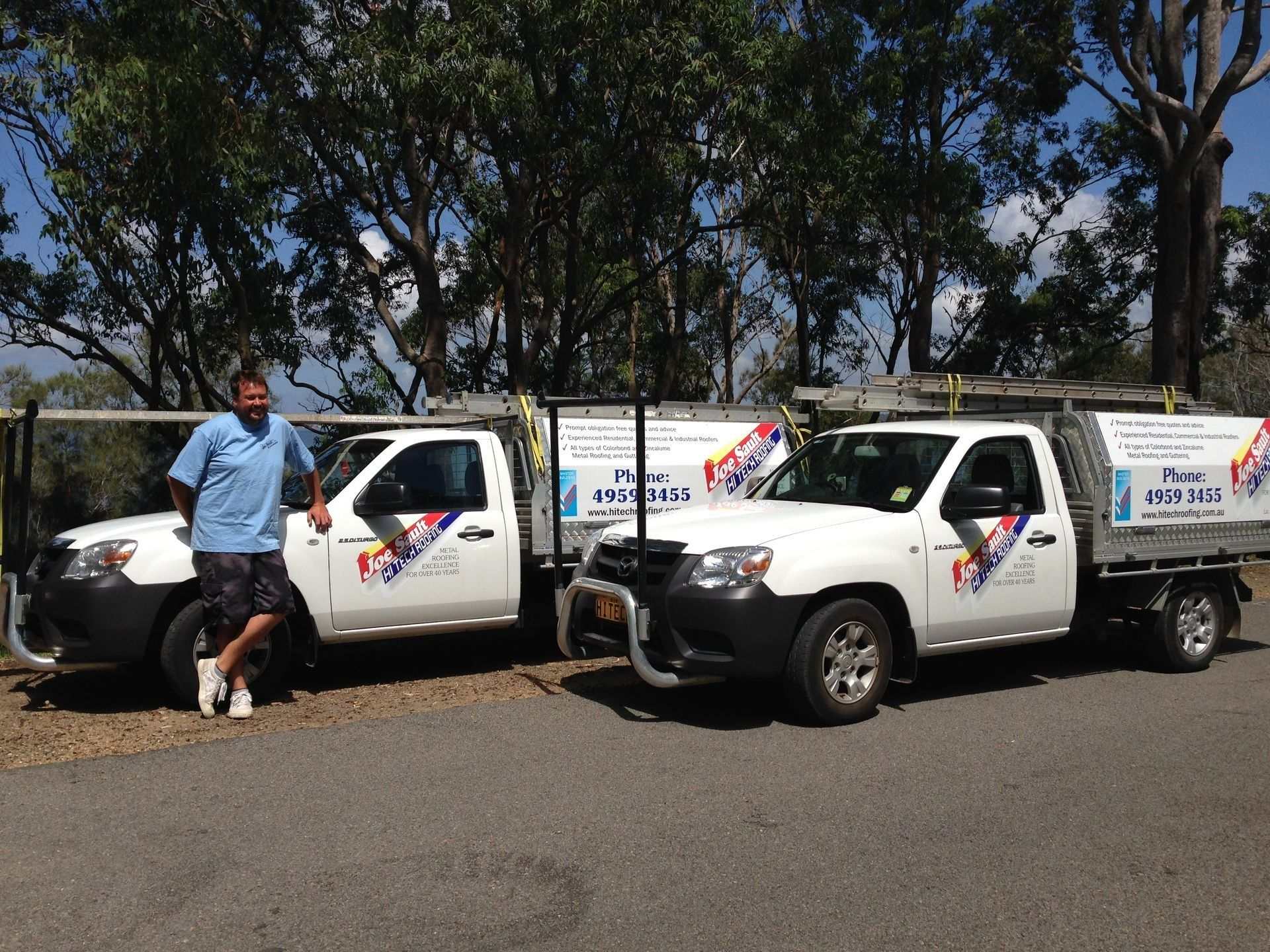 Man Stands by Two White Work Trucks with Company Logos; Sunny Outdoor Setting — Hi Tech Roofing in Kilaben Bay, NSW