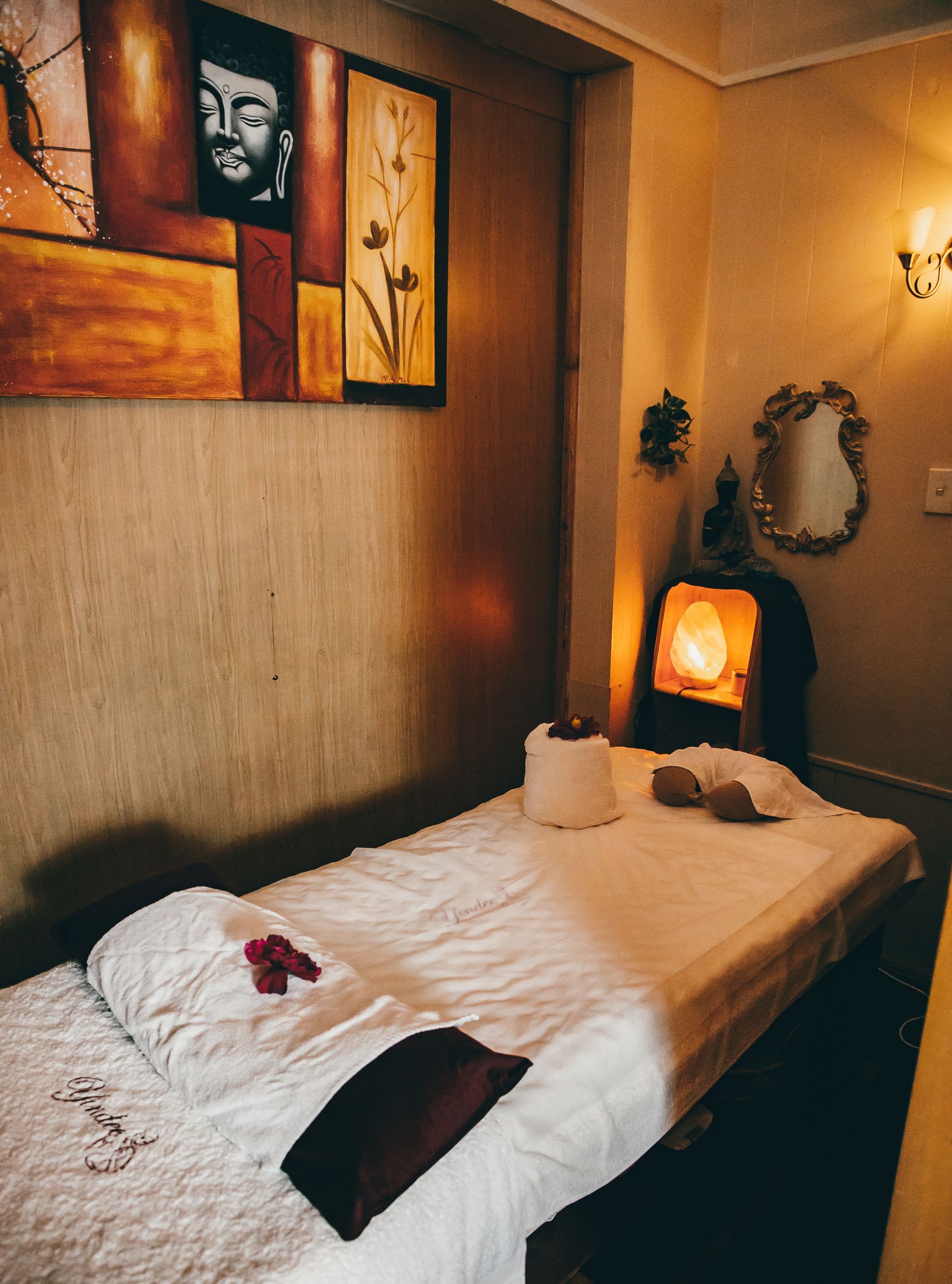 Massage room, bed with white linens, artwork on wall, warm lighting.