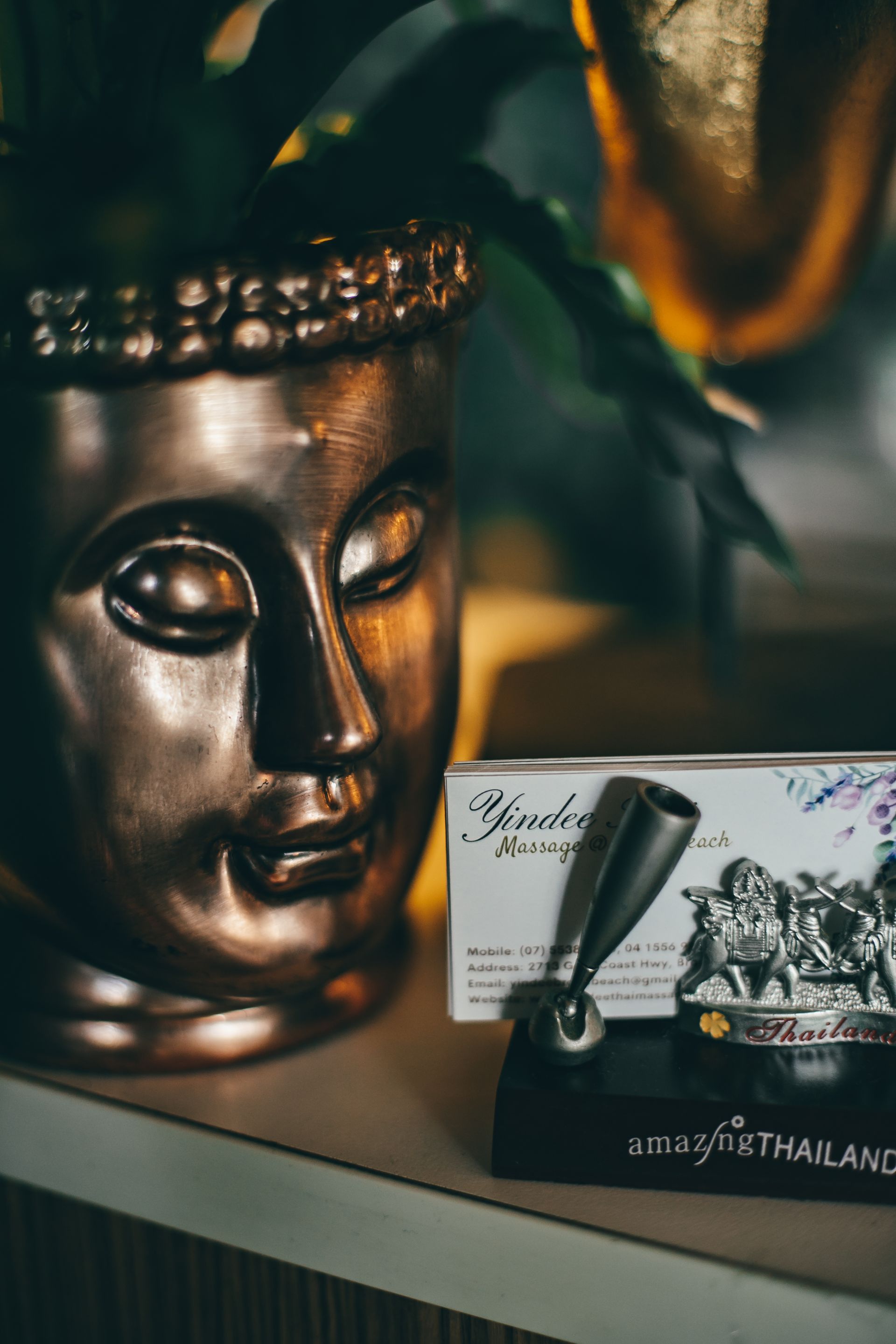 Bronze Buddha bust next to a pen holder on a shelf; green plant in the background.
