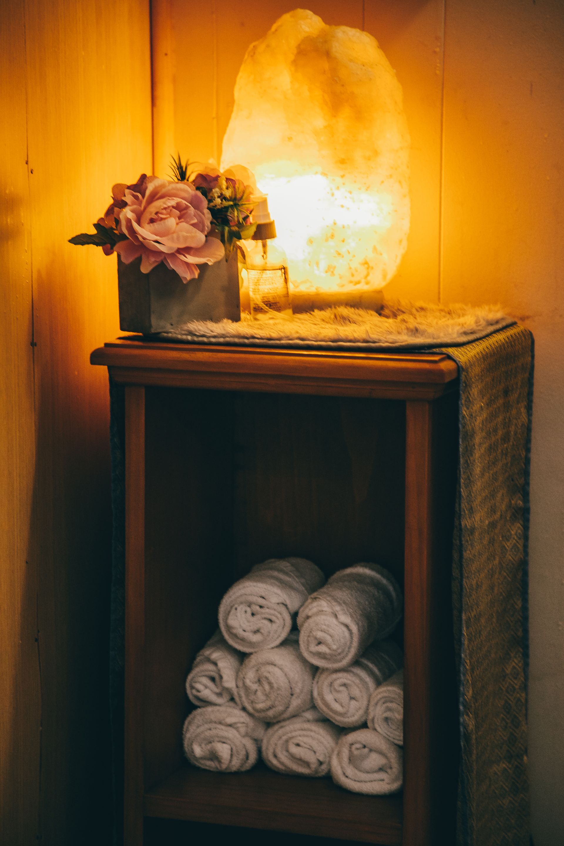 Wooden shelf with rolled white towels, a flower arrangement, and a glowing salt lamp.
