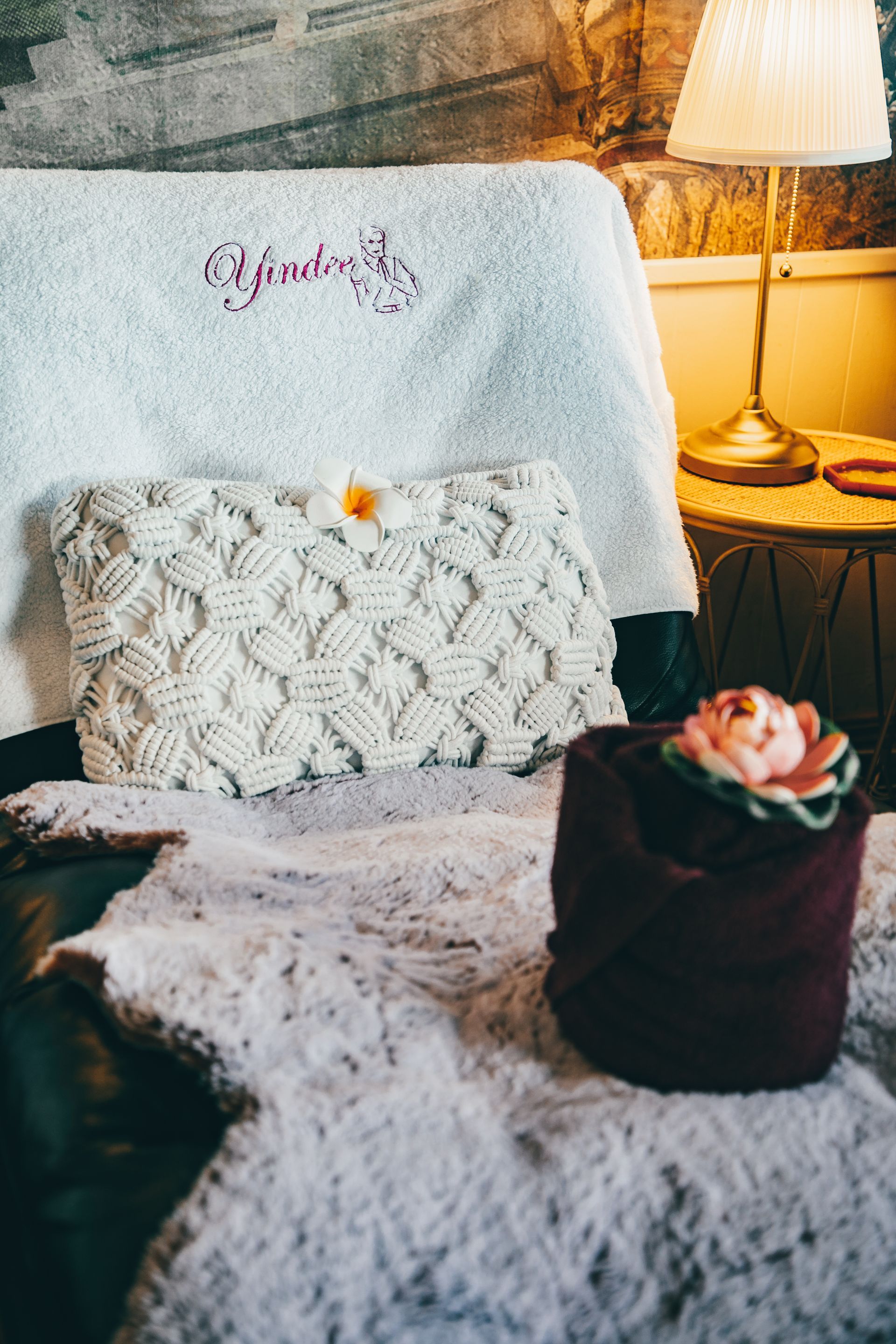 Cozy chair with a white woven pillow, towel, and a rolled towel topped with a pink rose in a warm-lit room.