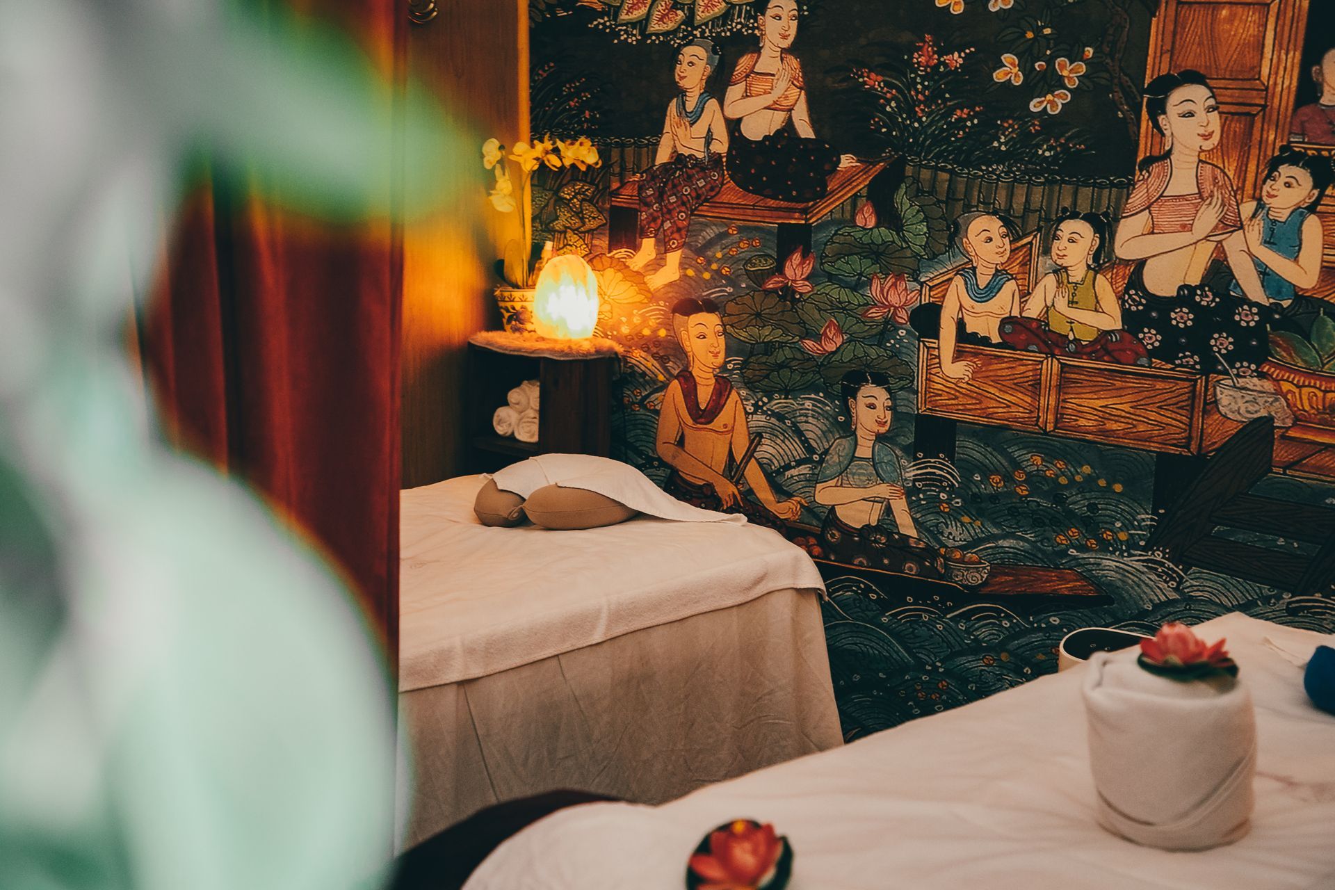 Massage room with two beds, floral decor, and a mural depicting figures. Soft lighting.