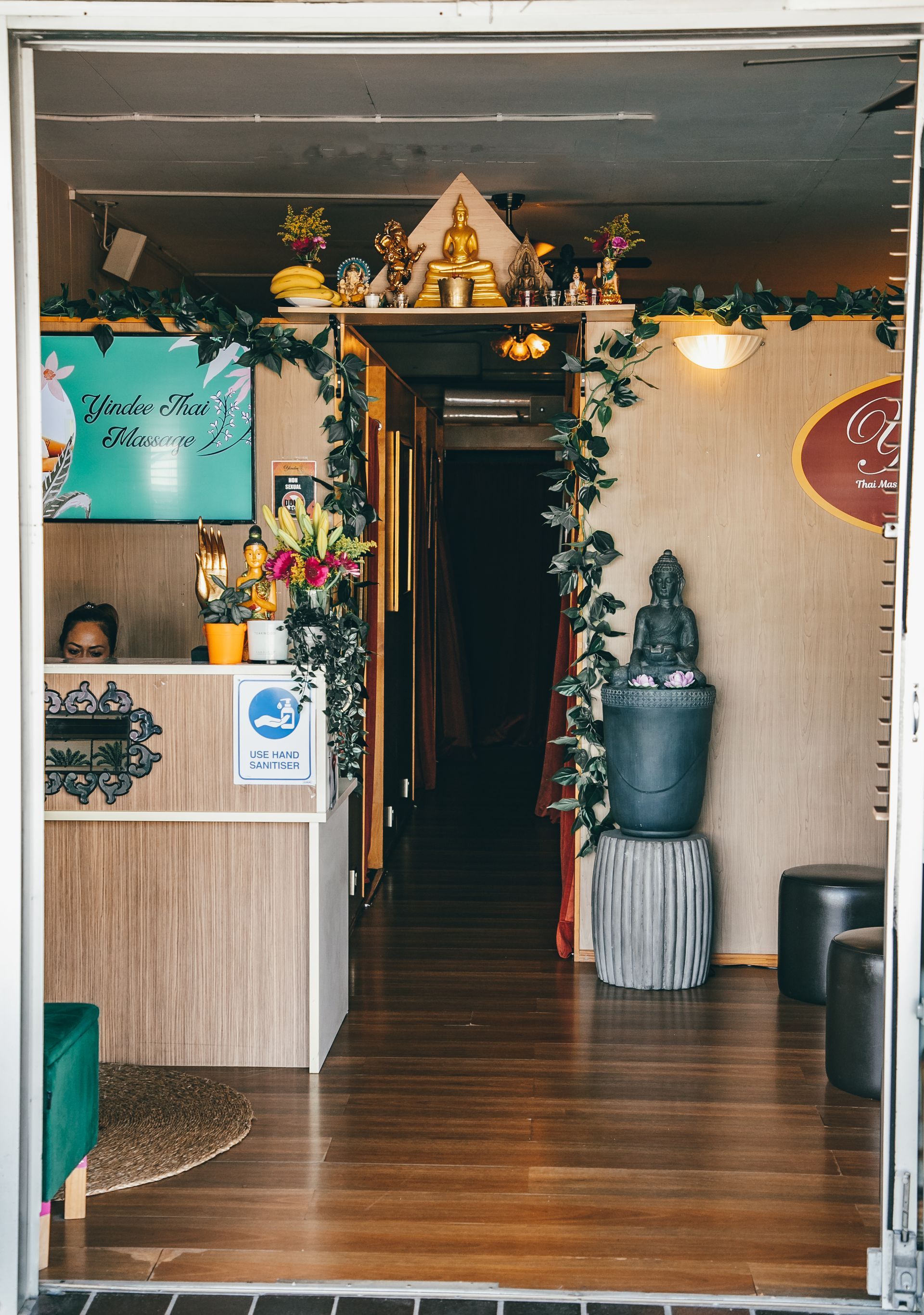 Entrance to a Thai spa, adorned with greenery and a small Buddha statue; receptionist at desk.