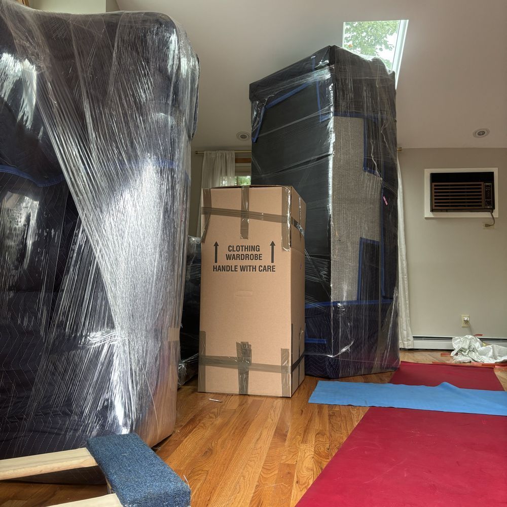 Furniture wrapped in plastic, cardboard box, blue and red floor runners, in a room.