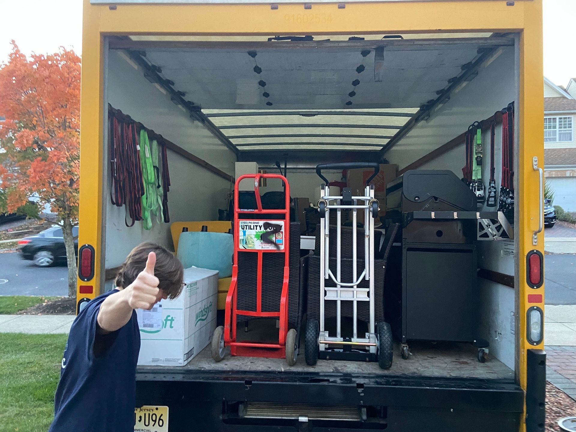Person giving thumbs up in front of a yellow moving truck. Truck bed contains boxes, carts, and a grill.