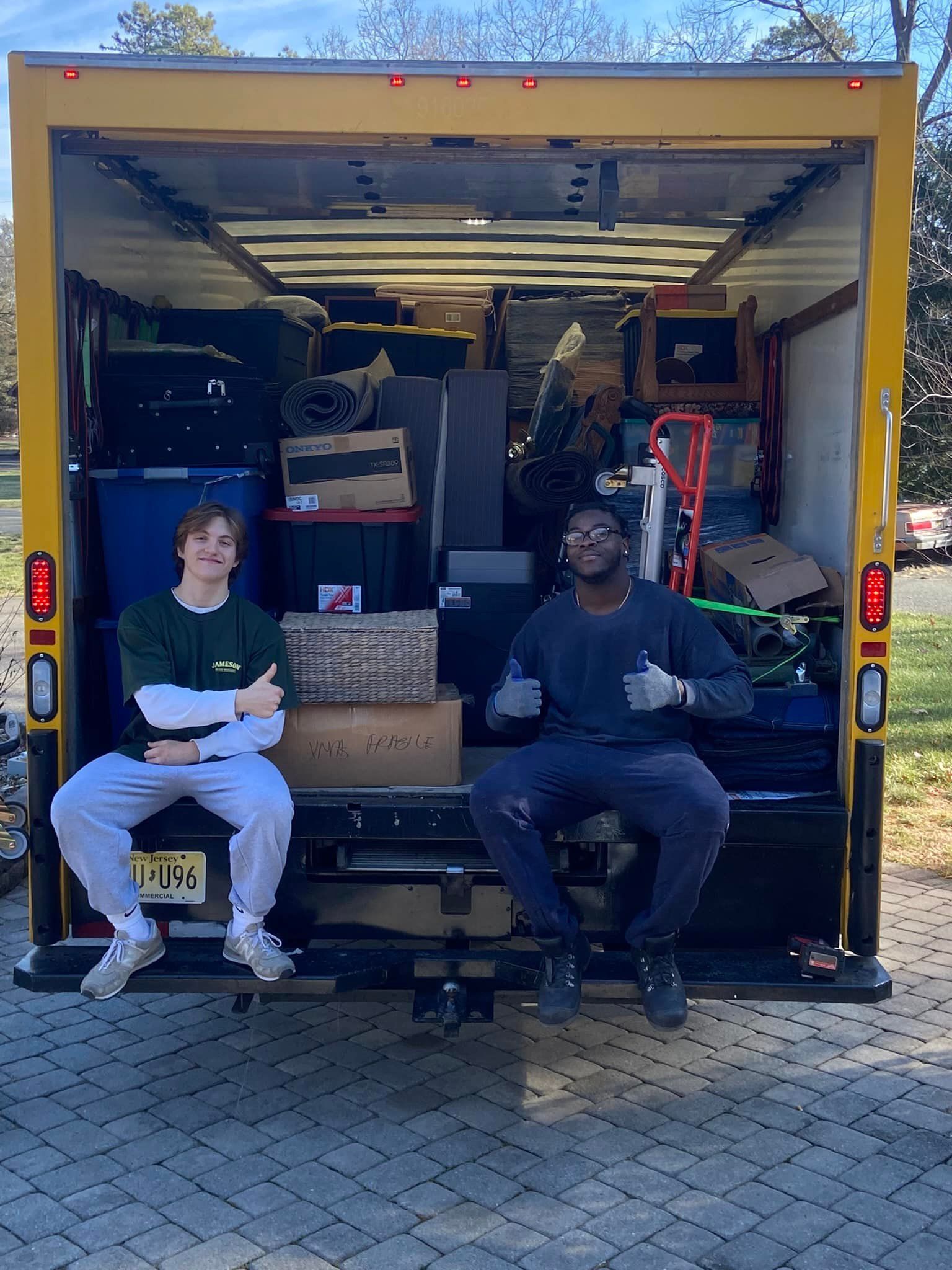 Two men sit in open moving truck. Truck is full of packed boxes. One man gives a thumbs up.