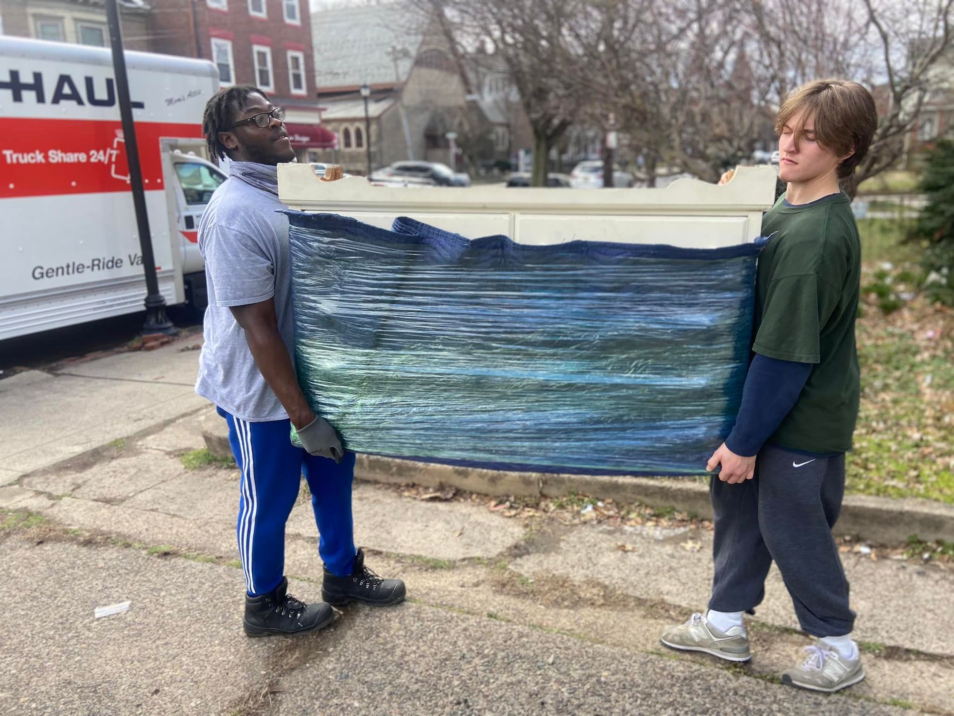 Two people carrying a large, wrapped rectangular object on a street, a U-Haul truck in the background.