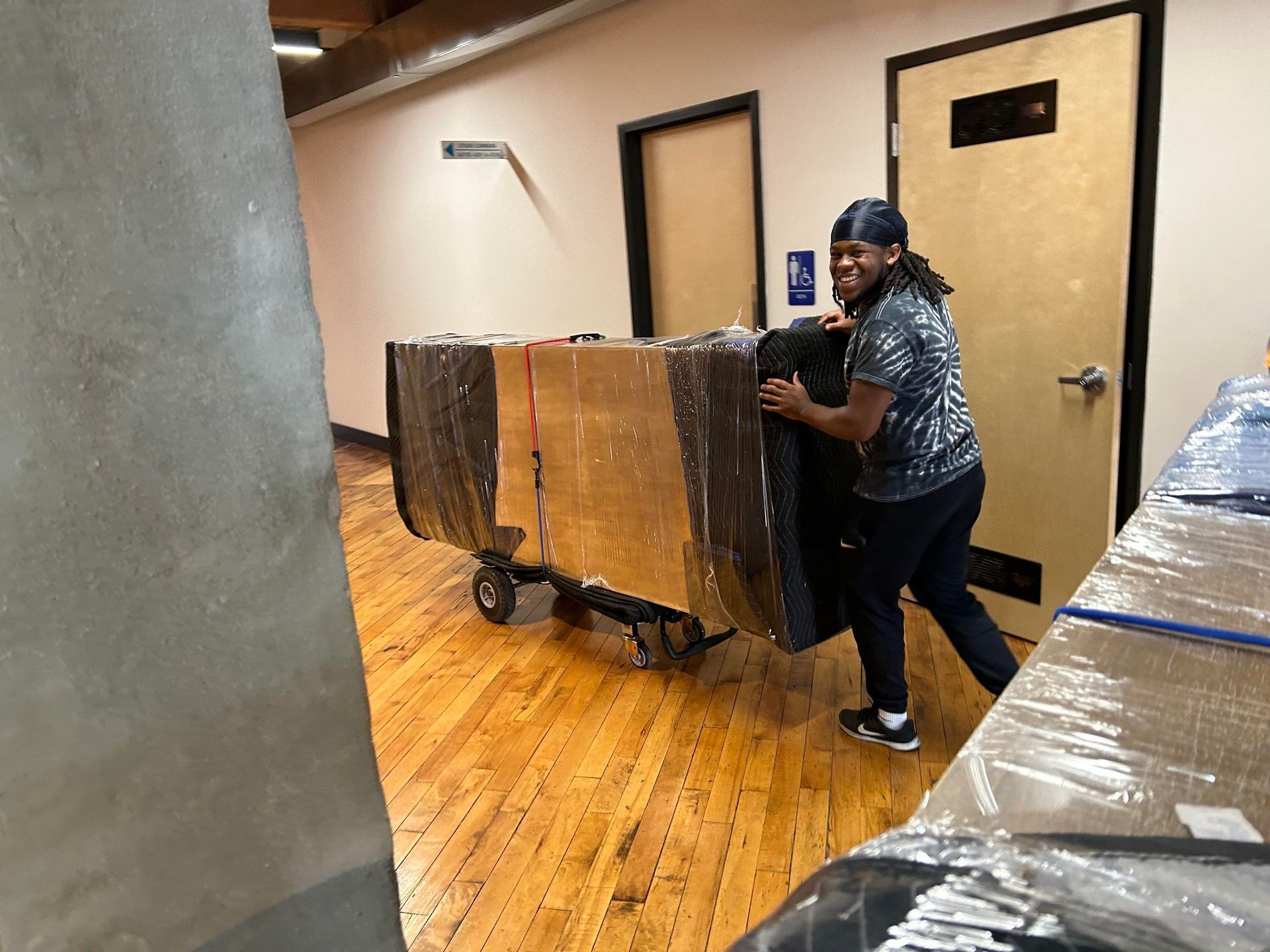 Person pushing a large, wrapped rectangular item on a dolly down a hallway.