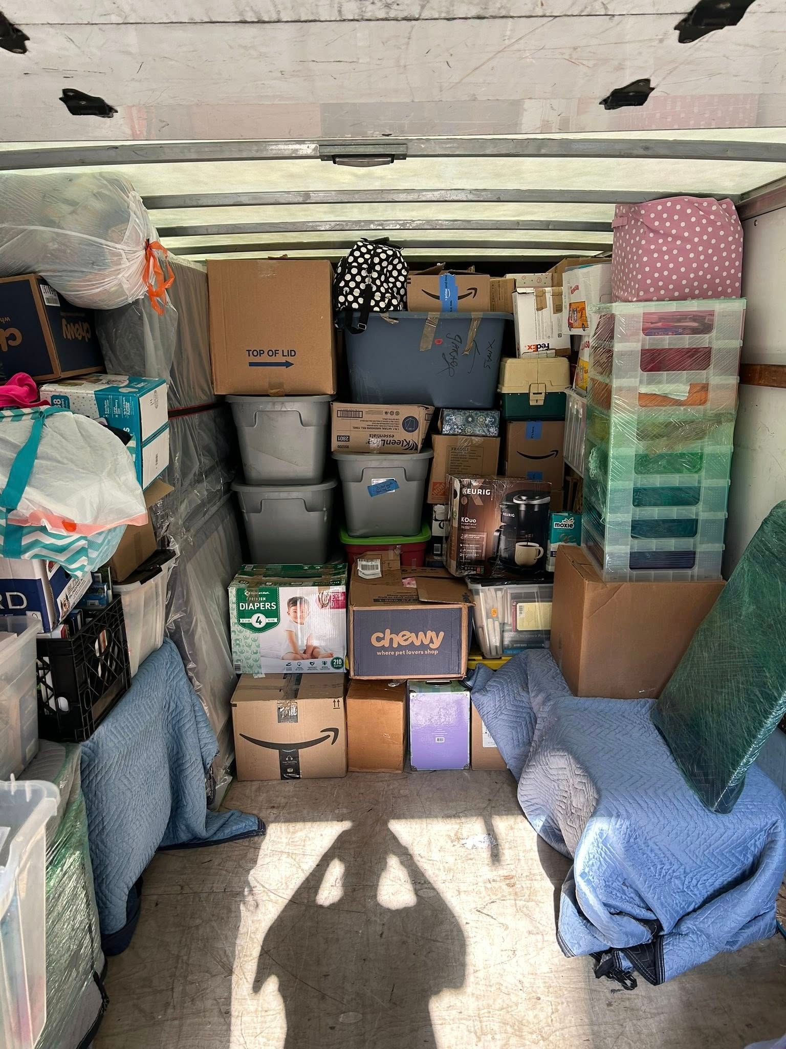 A packed moving truck interior: cardboard boxes, bins, and various items stacked, with a shadow of a person at the bottom.