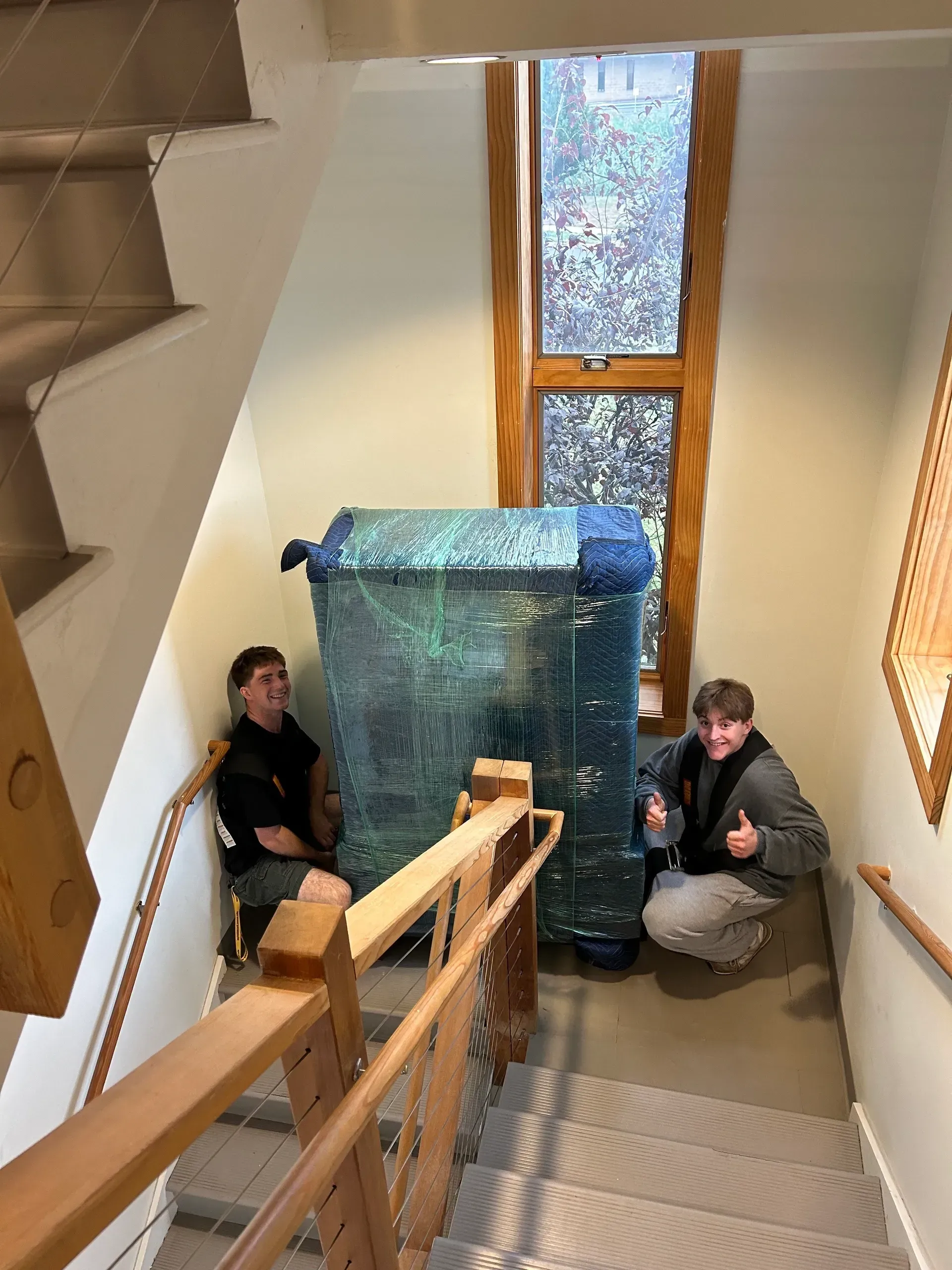 Two people on a staircase with a large, wrapped item. One gives a thumbs up.