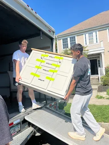 Two people lifting a white cabinet out of a moving truck on a ramp; sunny day.